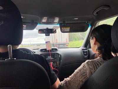 The image captures the interior of a car, viewed from the back seat. The front passenger seat is occupied by a woman with dark hair, wearing a beige knitted sweater and hoop earrings, looking out the window. The driver, whose back and arm are visible, is wearing a black long-sleeved shirt. The dashboard displays a clock showing 10:26. A phone mounted on a stand is visible, connected by a red charging cable. Outside the car, a truck is driving on a road, with other vehicles and greenery in the background. The scene suggests travel on a highway. The specific location in the city of Uzytamak, Russia, cannot be determined from the provided image alone.