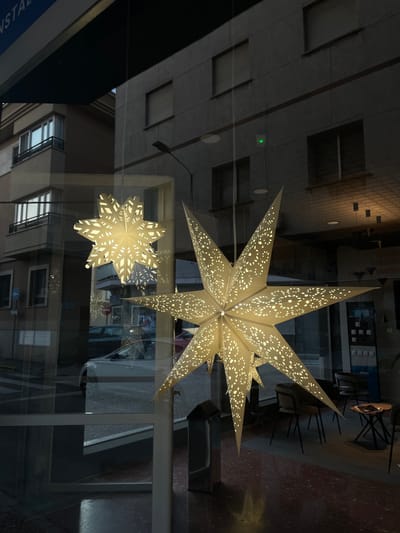 The image is a medium-angle shot of a storefront window on a street in La Línea de la Concepción, Spain. Two illuminated, paper star-shaped decorations are hanging in the window, casting intricate patterns of light onto the interior and reflecting the street scene outside. The larger star is in the foreground, partially obscuring the smaller star behind it.

Through the window, the exterior shows a gray, modern building with multiple stories and regularly spaced windows, some with closed blinds. A street with a crosswalk is visible, along with parked cars. The interior of the store is dimly lit, with a few chairs and tables visible, suggesting it might be a cafe or retail space. The floor inside has a speckled red and black pattern. A blue awning with white text, partially visible at the top of the image, indicates the establishment's signage. The overall impression is of a street-level commercial space, possibly decorated for a holiday.