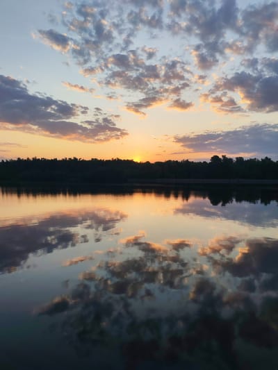 The image depicts a tranquil sunset scene at a body of water, possibly a river or lake, in Ermolino, Russia. The sky is filled with cumulus clouds tinged with orange and pink hues from the setting sun, which is partially obscured by a distant treeline. The surface of the water reflects the sky and clouds, creating a mirrored effect. The treeline is silhouetted against the bright sunset, providing a dark horizontal band across the middle of the frame. A faint contrail from an aircraft is visible in the upper center of the sky. The water appears calm with minimal ripples. The lighting suggests late evening or early morning.