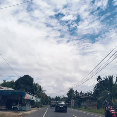 The image captures a rural or semi-rural road scene in Pandrahkandeh, Indonesia, under an overcast sky with patches of blue. The upper half of the image is dominated by the sky, filled with a mix of white and light blue clouds.
A two-lane asphalt road with white and yellow road markings stretches through the center of the frame, leading away from the viewer. In the mid-ground, a black SUV is visible from the rear, its license plate reading "BL 1330 AAF". Ahead of it, a light-colored truck, possibly a Mitsubishi, can be seen with "MITSUBISHI" printed on its tailgate. Both vehicles are moving away from the viewer. On the right side of the road, a red motorcycle with two occupants, one wearing a helmet, is driving towards the viewer.
The sides of the road are lined with lush tropical vegetation, including numerous tall palm trees and dense green foliage. On the left side, there's a simple, open-front roadside stall or shop with a blue tarp roof and a faded red, white, and blue banner. A smaller blue sign below the banner clearly displays the word "Rizki" and features an image of a truck. Further along the left, another building is partially obscured by trees. On the right, a wooden house with a red roof is visible amidst the trees. Multiple black power lines are strung across the sky, connecting utility poles on both sides of the road. The scene suggests a typical day with ongoing transit and local commerce in a tropical, rural setting.