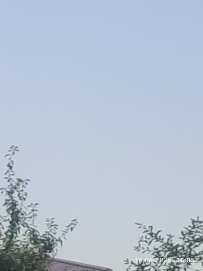 This media file is a vertical, blurry photograph of a clear, light blue sky. In the upper right corner, the out-of-focus leaves of a tree are visible, along with a sliver of a building with a purple-ish tint. Text indicating the date and time is superimposed vertically on the right side of the image: "16.06.2026 19:05". The time suggests it is late afternoon or early evening. There are no people or specific objects clearly discernible besides the tree and building. The overall scene is serene and outdoor-oriented, with the bright sky dominating the frame. The location context provided is Chighirik, Uzbekistan.