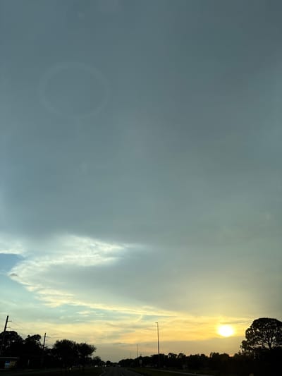 The image shows a rural highway in Venice, United States, during a sunset. The sky is partly cloudy with hues of yellow and orange, indicative of a sunset. A bright sun is visible on the right side of the frame, partially obscured by a large tree. The road stretches into the distance, lined with trees and utility poles on either side. There are no people or vehicles clearly visible on the road, suggesting a sparsely populated area or a moment of low traffic. The overall atmosphere is serene, with the natural beauty of the sunset dominating the scene.