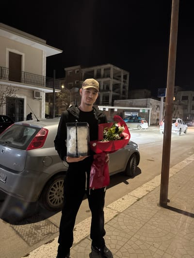 A young male stands on a tiled sidewalk in Stornara, Italy, at night. He is positioned facing the camera, slightly off-center to the left. He wears a beige patterned baseball cap, a black hooded sweatshirt, black sweatpants with white drawstrings, and black athletic shoes. He holds two items: in his left arm, a clear rectangular box containing several white rose-like objects, and in his right arm, a bouquet of red and white flowers wrapped in red material.

Behind him and to his left is a parked grey Fiat Punto hatchback, facing right. Further behind the man is a two-story building with light-colored stucco, a dark brown door on the upper level with a wrought iron balcony, and an air conditioning unit on the lower level. To the right of this building, a multi-story unfinished concrete structure is visible in the background, along with a smaller, low-rise building with a green awning. The street, partially visible behind the man, contains other vehicles and is illuminated by streetlights, casting ambient light on the scene. A dark utility pole is present on the right side of the foreground, just behind the man. The sky is dark, indicating nighttime.