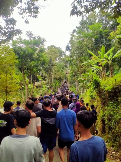 A large group of people walks up a narrow road surrounded by lush greenery. The road leads uphill, with trees and plants densely lining both sides. Most individuals are seen from behind, showcasing a variety of casual clothing styles. The sky is overcast, casting a soft light on the scene. The overall impression is one of a community gathering or pilgrimage in a rural setting.