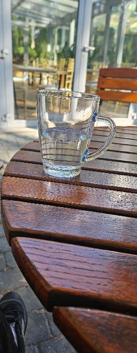 The scene is located in Mierzęcice, Poland. A clear, ribbed glass mug, containing clear liquid approximately halfway to the brim, stands centered on a dark brown, round wooden slatted table. The table surface is reflective, displaying highlights from a strong light source. To the immediate right of the mug, a segment of a wooden chair back, matching the table's material and color, is visible. The lower-left portion of the image shows the front of a dark athletic shoe with white accents and a dark pant leg, indicating a person seated at the table. The ground beneath the table consists of dark grey paving stones. The background features a large modern structure composed primarily of glass panels, resembling a conservatory or restaurant building. Through the glass, blurred interior elements including light-colored tables, chairs, and green foliage are discernible. The environment is brightly lit by natural sunlight, creating distinct reflections.