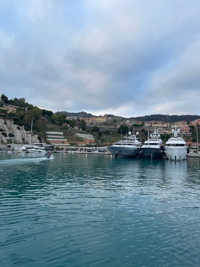 The image depicts a harbor in Ventimiglia, Italy, under an overcast sky. The foreground is dominated by deep blue water with visible ripples. In the mid-ground, several large yachts are moored. From left to right, there's a white sailing yacht, followed by three substantial motor yachts docked side-by-side. The motor yachts are modern and luxurious, with sleek white hulls and multiple decks. To the far right, another white yacht is partially visible.
A small inflatable boat with a single person in it is moving from left to right in the water, creating a noticeable wake. Behind the moored yachts, a waterfront promenade or dock area is visible, with some lighting fixtures and bollards.
The background showcases the city of Ventimiglia built on a hillside. Colorful buildings with terracotta roofs are clustered together, ascending the slope. The architecture suggests a Mediterranean style. Lush green trees and vegetation dot the hillside, interspersed with some grey rock formations and terraced areas. A construction crane is visible on the higher part of the hill. The overall atmosphere is calm and serene, with the overcast sky creating diffused lighting.