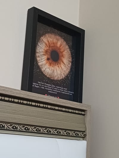 The image shows a framed print of a human iris. The iris is a warm, reddish-brown color with intricate patterns. Text is overlaid on the image, written in Bulgarian. The text reads: "Happy Birthday, my little heart! Years you are a part of me and my iris will always be with you, guarding our love and connection forever! I love you". The print is framed in a black, shallow box frame, and is partially obscured by a second, larger, ornate frame. This second frame has a light wood grain finish with decorative dark brown patterns featuring floral motifs. The overall impression is a personal gift, likely for a birthday, with a symbolic representation of connection through the iris. The setting appears to be indoors, possibly on a wall, as indicated by the neutral background and the way the frames are positioned. There are no indications of time of day or weather.
