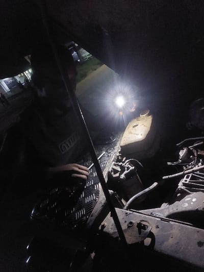 An adult individual, partially visible, is engaged in a nighttime vehicle repair in Msata, Tanzania. The scene depicts the open engine bay of a vehicle, illuminated by a bright, focused light source positioned towards the center-right. The individual, wearing a light-colored shirt with a "MTU" logo, has his right hand positioned over a section of diamond plate metal, where several small metallic objects, likely tools or parts, are visible.

Within the engine bay, a dirty, metallic engine block or related components are discernible on the right, along with various hoses and wires. A light-colored, soiled reservoir or tank is situated further back in the bay. A prominent crossbar runs horizontally across the upper portion of the engine bay. In the darkened background, faint lights from distant buildings or streetlights are visible, indicating an outdoor, possibly roadside or garage, urban setting.