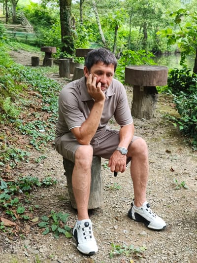 An adult male with short, dark hair and stubble is sitting on a log in a wooded area. He is wearing a brown polo shirt and matching shorts, a silver wristwatch, and white and black athletic shoes. His right hand is resting on his chin, and his eyes are closed. In the background, there are more logs that appear to be used as seating, a wooden staircase, dense green foliage, and a body of water. The ground is covered with gravel and fallen leaves. The scene is set in Kalinovoye ozero, Russia.