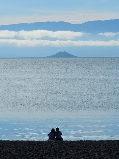The image depicts a landscape scene. In the foreground, there is a dark, textured shoreline. The central area is occupied by a body of water, showing subtle ripples. Beyond the water, the background presents a series of mountains in light blue hues, partially obscured by white cloud formations. The sky above is a clear, light blue. In the shoreline, two people are sitting next to each other. The location is near the city of Villarrica, Chile.