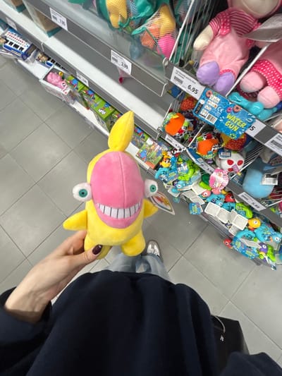 The image captures a first-person perspective of a person holding a distinct yellow and pink plush toy in what appears to be the toy aisle of a retail store. The plush toy, which is the central focus, is primarily yellow with a large pink section covering its face, featuring a wide, stitched smile with visible white teeth. It has two prominent, light blue googly eyes, one positioned above the pink facial area and another below. The person's right hand, with dark manicured nails, holds the toy.
The background reveals multiple metal-framed shelves stocked with various colorful toys. On the upper shelves to the left, pink plush toys are visible, along with multiple blue plastic "WATER GUN" packages. Further along the shelves, a variety of other toys are displayed, including colorful plastic items, some with eyes, and mesh bags containing smaller, brightly colored objects, possibly balls or toys. White pricing labels are affixed to the shelves, displaying prices such as "6.95", "5", "3.99", and "2.99". One visible label specifically mentions "WATER GUN" and the "CE" mark. Another small, pink tag partially visible behind other toys reads "SQUEAKY TOYS".
Below the shelves, the person's lower body is visible, dressed in dark long-sleeved clothing (possibly a hoodie or jacket) and light blue jeans. Their feet, clad in white sneakers, are also seen on the light grey tiled floor of the store. The lighting suggests an indoor retail environment. The image is taken from a slightly overhead angle and is rotated, giving a dynamic, casual snapshot feel.