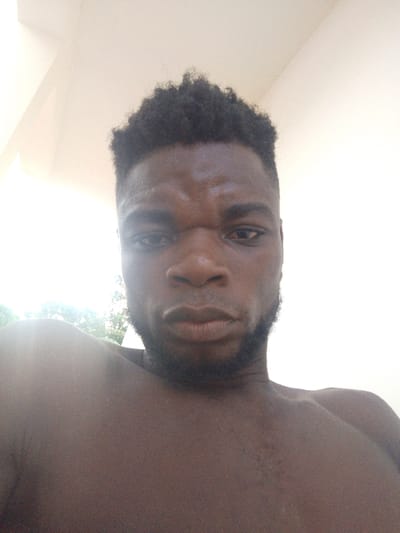 A close-up, low-angle shot of a dark-skinned man with a serious expression. He has short, dark, curly hair and a beard. His eyes are squinted, and his lips are pressed together. The man appears to be shirtless, revealing part of his shoulders and chest. The background is a blurred light color. The lighting is bright.