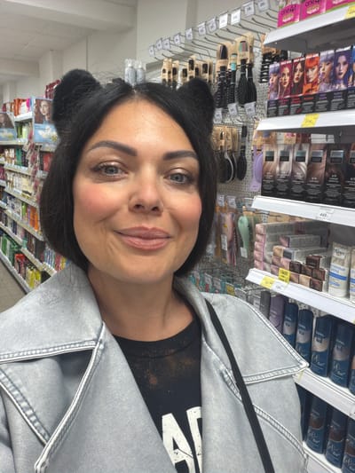 The image shows a woman wearing cat ears in what appears to be a store. She is wearing a silver jacket. The background shows store shelves stocked with products like hair dye and hairbrushes. The store appears to be a cosmetics or personal care aisle. The pricing tags indicate the location is likely in Russia, specifically in the city of Murmansk, as some tags have Russian words and currency symbols (rubles).