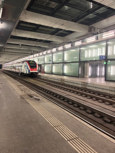 The image captures a modern train station platform, likely underground, in Chêne-Bourg, Switzerland. A sleek, multi-car train, predominantly white and red with blue accents on its nose, is stationary on the tracks. Its front end is visible in the distance, facing away from the viewer.
The platform, made of concrete or asphalt, is clean and features white tactile paving lines for the visually impaired. No people are visible on the platform or inside the train. On the left, a tall concrete structure with large, dark rectangular windows runs parallel to the platform, suggesting an adjacent building or wall.
The entire space is brightly illuminated by numerous overhead fluorescent lights and large, square, translucent ceiling panels that also appear to emit light, creating a well-lit, somewhat sterile environment. Above the tracks and platform, several signs are visible, including one prominent blue sign with "1 A" in white text, likely denoting a platform or track section. Green exit signs are also present. The train tracks are well-maintained, featuring concrete sleepers. The setting indicates a functional, contemporary public transport facility, potentially a metro or suburban rail line. The artificial lighting suggests an indoor or subterranean location, rendering the time of day and weather indeterminate.