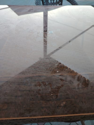 The image shows a close-up view of a wooden table with a glass top. Sunlight creates distinct shadows on the table's surface, indicating an outdoor setting. The wooden surface beneath the glass appears to be made of planks, with a weathered or textured finish. Some small debris or marks are visible on the table. In the background, out of focus, the edge of a tire or wheel and a portion of a wicker-like structure are visible, suggesting the presence of furniture or a vehicle nearby. The overall scene is likely a patio or outdoor seating area. The location is identified as Toranghyliq, Kazakhstan.