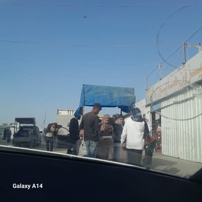 The image, taken from inside a vehicle (identified by the "Galaxy A14" watermark), shows a street scene in what is possibly the city of Khan Yunis. A group of people are gathered under a blue tarp-like structure that appears to be a makeshift shelter or covering. Some of them wear clothing that might include headscarves. There are vehicles, including what looks like a small truck, visible in the background and a building. The angle of the shot suggests the photographer is a passenger in the vehicle, which is moving.