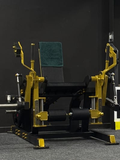 A piece of exercise equipment is situated against a wall. It's constructed from black and gold components, presenting a structured appearance. The machine has a seat with a dark-colored cushion and a green towel placed on it. Other elements include rollers and a handle, adding to its functionality. The machine is set up in what seems to be a home gym setting, with a dark background and a visible wall, implying an indoor workout space.