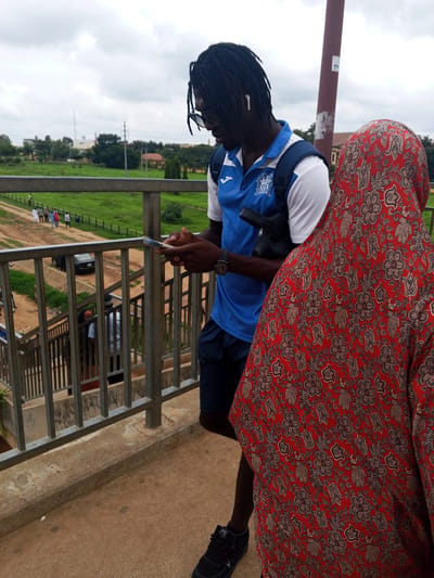 Two individuals stand on a concrete overpass with a metal railing. In the foreground, a person wearing a vibrant red garment with a floral pattern stands facing away from the camera. To their left and slightly behind, a person with dark dreadlocks, wearing a blue and white polo shirt and dark shorts, is looking down at their smartphone. They have a backpack and earbuds in. The overpass overlooks a grassy area with some buildings and trees in the background, suggesting an outdoor urban environment. The sky is overcast. There are other people walking on a path below the overpass. The location is identified as Immigration Quarters, Nigeria.