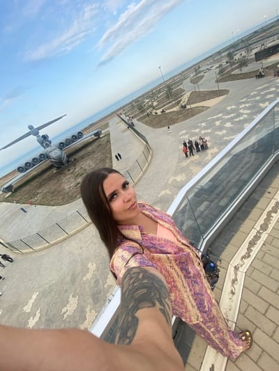 A high-angle, wide shot captures a woman in the foreground, her arm extending into the frame holding the camera. She is wearing a vibrant, patterned outfit in shades of pink, purple, and yellow, and appears to be standing on a paved area with a glass railing. Behind her, a large, gray airplane rests on a grassy, dry field, its numerous engines visible. The plane is positioned near a body of water, likely the Caspian Sea, given the presumed location of Arablyar, Russia. Beyond the plane, a park area with walking paths, benches, and scattered trees is visible, with a group of people gathered in the middle distance. The sky is a clear blue with sparse, wispy clouds. The overall scene suggests an outdoor exhibition or park setting with a historical aircraft as a centerpiece.
