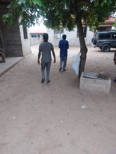 The image depicts an unpaved outdoor area in Mwandege, Tanzania. Two individuals are walking away from the camera. The lead individual, a male, wears a black and white striped short-sleeve shirt, grey trousers, and dark flip-flops, with two bracelets on his left wrist. The second individual, also male, walks to the right of the first, wearing a blue long-sleeve shirt, grey trousers, and dark sandals. A third person's lower legs, clad in blue pants and dark shoes, are partially visible at the far right edge of the frame. The ground is light brown, compacted dirt with scattered small debris. A prominent tree with dense green foliage stands right of center; a white plastic bag is tied to its trunk or a low branch. A rectangular concrete block with a dark, irregularly shaped object resting on top is situated on the ground near the tree's base. In the background, a building constructed from light-colored concrete blocks features multiple windows, some appearing shuttered. To the right, a dark-colored Land Rover Defender-style vehicle is parked with a spare tire mounted on its rear. Another building of similar construction with a window is partially visible on the far left. The sky overhead appears light and overcast.