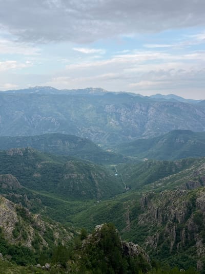 The image depicts a panoramic view of a lush, mountainous landscape under a cloudy sky. The foreground features rugged, rocky terrain covered with sparse green vegetation and a few trees. As the view recedes, progressively layered mountain ranges are visible, cloaked in dense green forests. A narrow, winding river or road can be seen cutting through the valley in the mid-ground, leading towards a small, tranquil body of water. The distant mountains are softened by atmospheric haze, appearing in shades of blue and grey. The scene conveys a sense of natural grandeur and remote wilderness. There are no people or objects that would indicate a specific location within the city of Aranđelovo, Bosnia and Herzegovina. The environment is entirely natural and undeveloped.