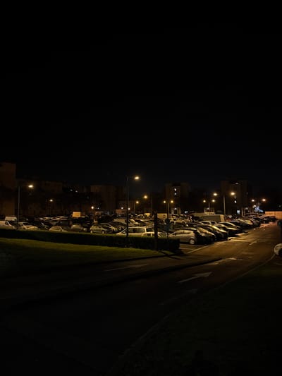 A dimly lit nighttime scene depicts a parking lot filled with cars along the right side, illuminated by streetlights. Adjacent to the parking lot is a road marked with a white arrow, bordered by a patch of grass. In the background, buildings with lit windows are visible, receding into the distance. The left side of the frame is dominated by a large expanse of dark sky.