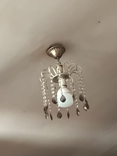 A ceiling light fixture with faux crystal drops hangs from a pale ceiling in Alagyaz, Armenia. The fixture has a metallic finish and is illuminated by a single, partially visible lightbulb. The ceiling appears to be textured and slightly discolored in places, with a few small marks or imperfections visible. The overall impression is of an indoor domestic setting.