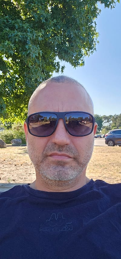 A man with a shaved head and a gray beard is pictured from the chest up, wearing black sunglasses and a dark blue t-shirt. The sunglasses reflect trees, a building with a tower, and several parked cars. The man is outdoors, with a large tree to his left and a clear blue sky above. The background shows a grassy area with a few large rocks and more trees in the distance, suggesting a park or rural setting. A grey SUV is parked on the right side of the frame. The setting is consistent with the town of Cestas, France, known for its forested areas and outdoor recreational spaces.
