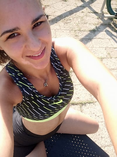 The image depicts a young adult female in an outdoor setting within the city of San Marzano sul Sarno, Italy. She has a light complexion, dark hair pulled back, and dark eyes. A noticeable light-colored patch is present on her upper lip, philtrum, and chin area. She is smiling and looking directly at the camera, appearing to take a self-portrait with her left arm extended. Her attire includes a black sports bra with a lime green bottom band and a white and lime green abstract pattern, displaying a white Nike swoosh logo on the left chest. She is also wearing dark shorts. A silver-colored necklace with a small pendant is visible around her neck. She is positioned as if seated or kneeling on a dark, possibly textured mat. The ground in the background consists of dry, light-colored soil or gravel with sparse dry grass. Strong, defined shadows are present, indicating bright sunlight. A portion of a metal structure with a green component is partially visible in the upper right background.