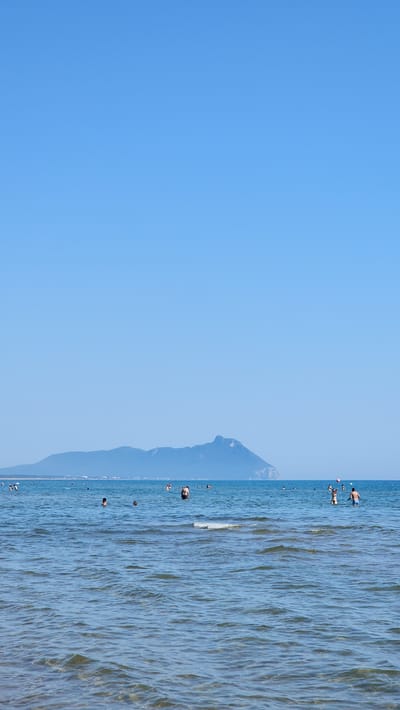In Sabaudia, Italy, a clear, bright sky dominates the upper portion of the image. Below the sky, a tranquil blue sea stretches out, dotted with numerous people enjoying the water. In the distance, a long, mountainous island or coastline forms a hazy, blue silhouette against the horizon. The water is calm, with gentle ripples reflecting the sunlight. Several individuals are visible swimming or standing in the shallow areas of the sea.