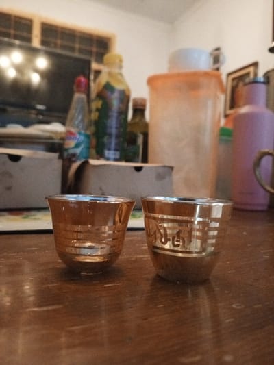 The image presents an indoor scene in Longmiang, Laos, featuring two decorative gold-colored cups on a dark brown wooden table in the foreground. The cup on the left exhibits several horizontal gold bands, while the cup on the right displays similar bands along with stylized gold Arabic calligraphy on its side. Both cups possess reflective silver interiors. Behind the cups, a partial view of a white and green patterned mat is visible on the table. The background is out of focus, revealing a cluttered shelf or surface. Notable objects in the blurred background include a television screen reflecting multiple light sources, a window frame with horizontal wooden slats, a slender clear bottle with a red cap, a taller clear bottle with a yellow cap, a dark glass bottle, a large opaque light orange plastic container with a white lid, a light pink bottle, and a partial view of a gold-colored handle from another vessel. The wall behind these items is light-colored. No individuals are present, and no direct actions or interactions are depicted.
