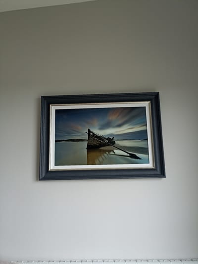 A framed artwork hangs on a plain, light grey wall. The artwork depicts a beached, decaying wooden boat on a sandy shore, with a calm body of water in the background. The sky above shows streaks of blurred clouds, suggesting a long exposure photograph. The frame of the artwork is dark grey with a silver inner mat. Below the artwork, a white radiator with horizontal bars is visible. The image is taken from a slightly angled, low perspective, looking up at the artwork. The location is specified as Doirí Beaga, Ireland, though no specific landmarks or features within the image confirm this directly. There are no people or animals present in the scene.