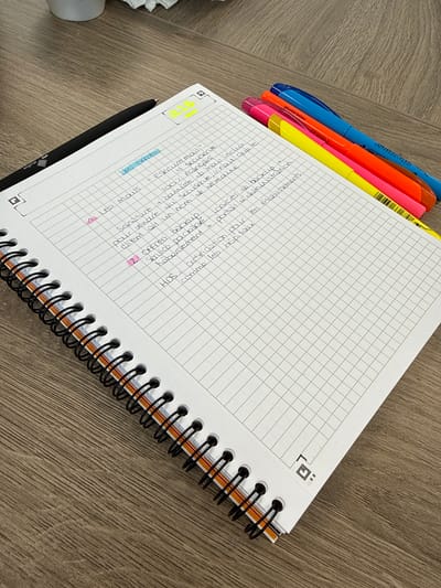 A close-up, eye-level shot shows a notebook on a wooden desk. The notebook is spiral-bound with a white cover and grid paper. Several colored highlighters and a black pen lie on the notebook, along with handwritten notes. The notes are in French and discuss items like "Les mails," "signature," "backup," and "certification." The background is blurred, showing a white object and a crumpled piece of paper. The scene is indoors, likely a desk setup in an office or study, and there are no indications of the location being in Les Arcs, France.