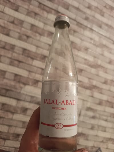 A person's hand holds a clear glass bottle of "JALAL-ABAD" mineral water. The bottle has a white label with red and silver text. The label prominently features the brand name "JALAL-ABAD" in large red letters, followed by "KELECHEK" in smaller red letters. Below that, in a script font, is text that appears to be Russian, and the number "27" within a red circle. At the bottom of the visible label, "ГАЗИРОВАННАЯ" is written in red. The background is a blurred pattern of horizontally oriented beige and white bricks. The lighting suggests an indoor setting, possibly during the evening. No specific activity or event is discernible, other than the act of holding the bottle.