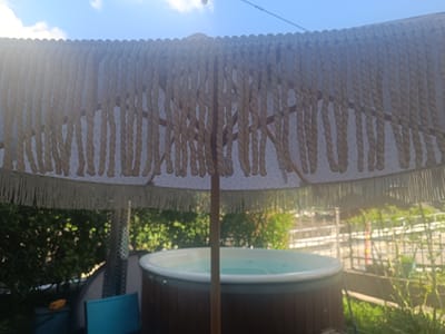 A low-angle, medium shot captures a close-up view of a thatched umbrella's fringe, obscuring a portion of the sky and background. The fringe is composed of numerous beige ropes, each intricately twisted, hanging in dense rows. Beneath the umbrella, a circular hot tub with a wooden exterior is visible, filled with clear blue water. The background features lush green foliage and hints of outdoor structures, bathed in bright, diffused sunlight. The scene appears to be an outdoor recreational area, possibly a backyard or patio, in Serravalle, Switzerland. There are no people visible in the image.