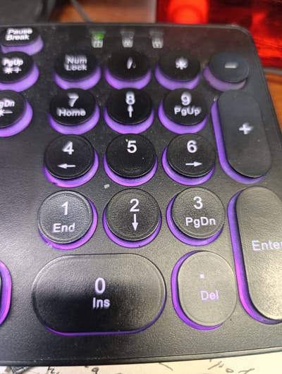 The image shows a close-up of a black keyboard with purple backlighting. The keys are round and black with white text. Visible keys include "Pause/Break", "PgUp", "Num Lock", a period, a star symbol, a dash, "PgDn", "Home", "8" with an up arrow, "9", "4" with a left arrow, "5", "6" with a right arrow, "1" with "End", "2" with a down arrow, "3" with "PgDn", "0" with "Ins", "Del", and "Enter", as well as a plus key. The keyboard appears to be in a dimly lit indoor setting, possibly a desk. There is no indication of Moytevo, Russia, or any people or specific actions.