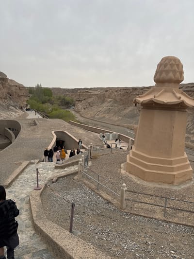 The image depicts a tourist area within the city of Guazhou County, China. A stone structure resembling a column or monument with a decorative pineapple-like top is prominent in the foreground. A pathway descends into an area with cave entrances, where a group of people is visible, suggesting a cave exploration site. The surrounding environment is characterized by rocky cliffs and sparse vegetation. The sky appears overcast, indicating possibly inclement weather. Some individuals are walking on the path, and others are gathered around the cave entrances. Railings are in place along the pathway to guide and protect visitors.