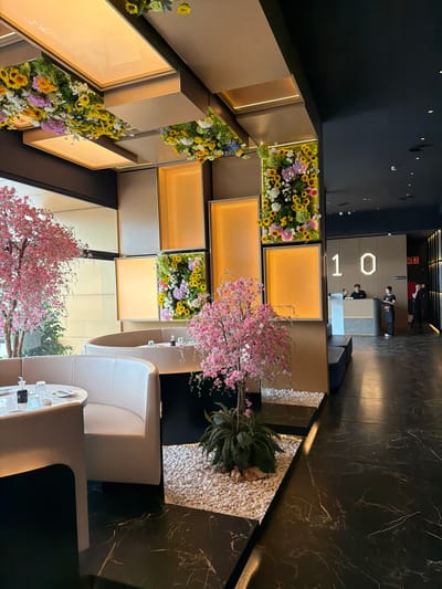 The image shows an interior space with a decorative wall featuring flowers and illuminated panels. A reception area with personnel is visible in the background, displaying the number "101". The foreground includes seating arrangements with white chairs and tables, adorned with artificial cherry blossom trees and a decorative rock garden with ferns. The environment suggests a modern or upscale establishment. There is no indication this is in Torreforta, Spain.