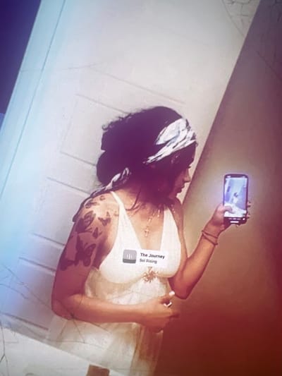 A woman is holding a smartphone in her right hand, seemingly taking a selfie or recording a video. She is wearing a white top with thin straps and a bandana on her head. Her left arm has tattoos of butterflies. The background appears to be an indoor setting with a wall and a door. The lighting is dim and has a reddish-purple hue, possibly due to filters or editing. There is a music player interface visible on the smartphone screen, displaying "The Journey Sol Rising." No specific location in Boone, United States of America, can be identified from the image.