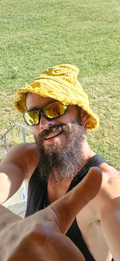 A man is pictured outdoors on a sunny day, wearing a yellow bucket hat and sunglasses, with a long beard. He is smiling and giving a thumbs-up gesture towards the camera. The background consists of a grassy area and a railing. The image appears to be taken in Wattenwil, Switzerland, likely during a warm season.