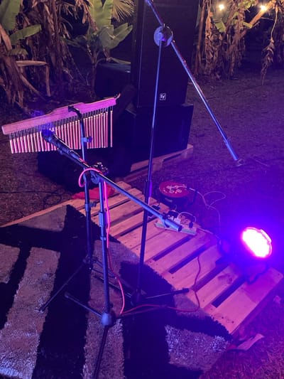 The image depicts an outdoor musical performance setup at night in Murera ward, Kenya. The scene is illuminated by artificial purple-pink lighting.

In the foreground, a black and grey patterned rug lies on grassy ground. Two microphone stands are positioned on the rug. One stand supports a microphone aimed at a set of wooden-topped bar chimes with metallic tubes. A second boom microphone stand extends diagonally across the frame. Red audio cables are visible on the ground and wrapped around the stands.

In the midground, a stack of two black "EV" (Electro-Voice) branded speaker cabinets rests on wooden pallets. To the right, another wooden pallet serves as a platform for a red power reel and a multi-outlet power strip with various cables connected. Adjacent to these power components, a round LED stage light emits a bright purple-pink beam.

The background features dense, dark foliage, consistent with banana plants or similar large-leaved vegetation. Faint, warm-white string lights are visible strung among the trees in the upper right portion of the background, contributing to the ambient lighting. No individuals are present in the scene.