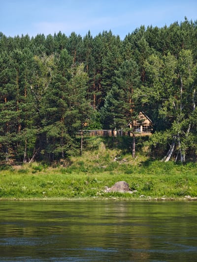 The image depicts a tranquil scene on a riverbank in Podporozh'e, Russia, under a clear blue sky. In the foreground, a wide river with calm, green-tinted water reflects the surrounding environment. The middle ground features a steep, grassy embankment covered in lush vegetation, with a cluster of large rocks near the water's edge. Higher up on the embankment, nestled amongst a dense forest of pine and birch trees, is a small, wooden cabin with large windows. The forest extends to the horizon, creating a verdant backdrop. The cabin appears to be a secluded dwelling, possibly a dacha, surrounded by the natural beauty of the Russian wilderness. There are no people or animals visible in the frame.