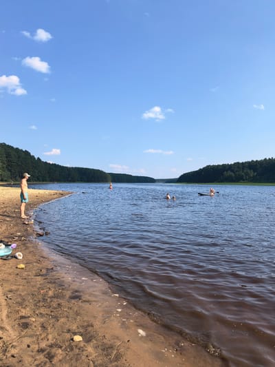 This media file depicts a serene summer day at a lake in Sysert, Russia. The scene is dominated by a wide expanse of calm, dark water, reflecting the bright blue sky dotted with fluffy white clouds. A sandy shore in the foreground is lined with scattered rocks and some discarded footwear, including flip-flops and sandals, suggesting recent human activity.
On the left side of the frame, a man wearing blue swim trunks and a white cap stands on the beach, looking out at the water. He appears relaxed, enjoying the view.
Further out in the lake, several people are engaged in recreational activities. Two individuals are swimming, while another person is on a stand-up paddleboard. A bright orange buoy is visible in the distance, marking a point in the water.
The background is a lush, dense forest that stretches along the far bank of the lake, creating a natural and tranquil setting. The overall impression is one of a peaceful summer day, with people enjoying the water and the natural beauty of the Russian landscape. The time of day appears to be late afternoon or early evening, given the soft lighting. There is no visible text in the media file.