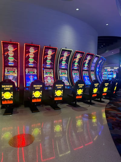 The image depicts a brightly lit, unoccupied casino gaming area, likely located within a casino in Paradise, United States. A prominent, curved row of at least nine "Dragon Link" slot machines is positioned from the left foreground extending towards the right background. Each machine features a large, vertical screen displaying vibrant game graphics, including jackpot progressives labeled "GRAND," "MAJOR," and "MINOR," along with Asian-themed characters such as a Buddha figure and warriors. Visible jackpot amounts include $19,470.98, $18,878.08, and $18,870.08. The slot machines are framed with integrated red LED lighting.
In front of each slot machine is a black gaming chair, with the backrest displaying an illuminated "DRAGON LINK" logo and a stylized golden orb over a fiery orange base. The polished light-colored floor in the foreground is highly reflective, showing clear reflections of the machines' and chairs' illumination. Towards the right, the floor transitions to a section of multi-colored patterned carpet. The background features a tall, curved wall with a textured surface, illuminated by ambient blue light from overhead recessed fixtures in the ceiling. No individuals are present within the scene, and no actions or interactions are depicted.