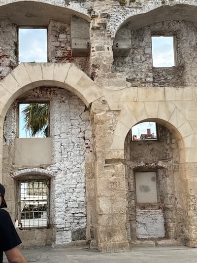 The image captures a section of the ancient Roman Diocletian's Palace in Split, Croatia, focusing on the weathered stone walls and arched openings of the structure. The stone is a mix of rough-hewn blocks and smoother, dressed stones forming the arches and some of the walls. The masonry shows signs of age and decay, with exposed brickwork and crumbling plaster in many areas.
Several windows are visible within the stone walls. One large, rectangular opening at the top left frames a cloudy sky. Below it, a significant arched opening reveals a window with a metal grate, through which a palm tree is visible against a bright blue sky. To the right, a smaller, higher arched window looks out onto rooftops with red tiles and an antenna. Further down and to the right, another arched opening contains a boarded-up window.
The foreground shows a paved walkway, with a partial view of a person on the far left, wearing a black t-shirt and a black baseball cap. They are facing away from the camera, and only their shoulder and side of their head are visible. The lighting suggests it is daytime, with the sun casting some shadows. The overall impression is one of historical preservation and the enduring presence of ancient architecture within a modern city.