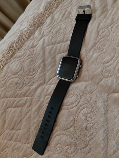 A black smartwatch with a silver buckle and casing is positioned diagonally on a plush, light brown quilted fabric. The fabric features a subtle swirling pattern. The smartwatch's black strap has several adjustment holes visible. The image was taken indoors, likely in Preobrazhenskoye, Russia.