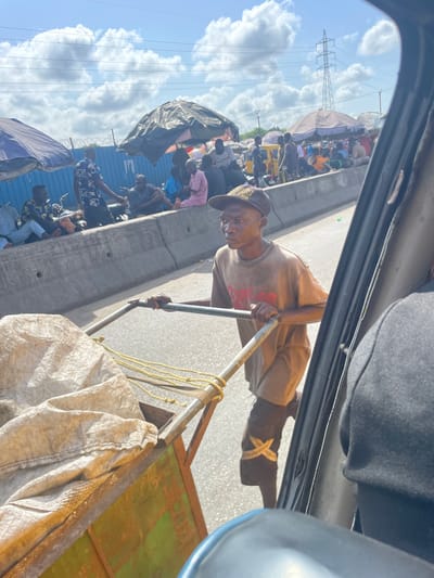 The image, taken from inside a vehicle looking out a window, captures a vibrant street scene in Alasia, Nigeria, under bright daylight with scattered white clouds in a blue sky.
In the immediate foreground, the interior of the vehicle is visible. Just outside, a man is seen pushing a metal cart along a paved surface, likely a road. He is wearing a light brown t-shirt, a dark cap, and dark boots, one of which features a distinctive light-colored "X" design. The man looks directly towards the camera with a neutral, perhaps focused, expression. His cart carries items, including a large white, possibly fabric, bag and yellow ropes, with a greenish-brown base visible.
A concrete barrier runs parallel to the road, separating the man from a densely populated area beyond. This area is bustling with numerous people, some standing, many gathered or seated under various makeshift shelters and umbrellas, suggesting a busy market or roadside vending area. Tarps and tents are used to create these temporary structures, indicating commercial activity.
In the background, beyond the crowded market area, a tall metal power pylon stands prominently against the sky. The overall scene depicts a dynamic outdoor environment, filled with human activity and daily commerce under clear, sunny conditions. No clearly legible text is visible on clothing or signs.