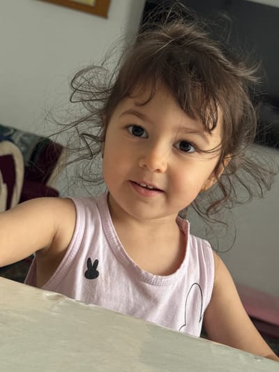 A young child with brown hair and dark eyes is looking at the camera. The child is wearing a pink tank top with a black bunny silhouette. The child is sitting at a table, with the camera angled slightly upwards. The background is blurred, but appears to be an indoor setting. There is no indication of the city of Ləcət, Azerbaijan in the image.