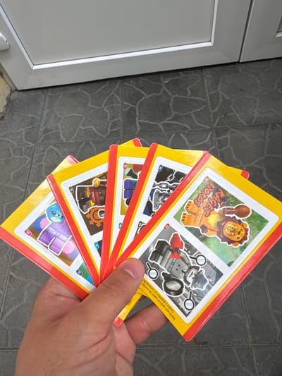 A person's hand holds a fan of several colorful trading cards, each featuring a Lego minifigure or character. The cards have yellow borders with red trim and white sections containing images and numbers. The background is a gray tiled floor and a white door. The scene is outdoors.
