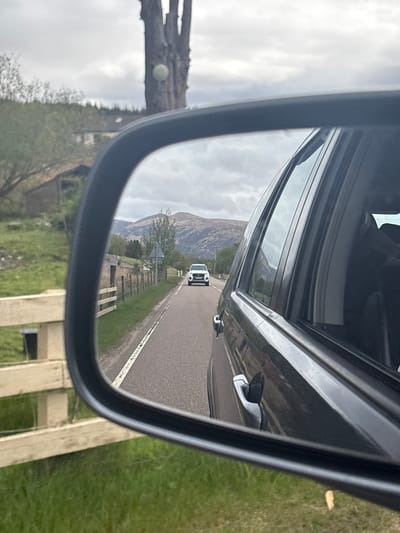 The image depicts a view from a car's side mirror, showing a road receding into the distance. A white car is visible on the road behind the vehicle. The surrounding environment includes green grass, a stone fence, and a mountain in the background. Part of a stone monument is reflected in the mirror as well, due to the location being in the Spean Bridge, United Kingdom area. The image is taken during daytime, with overcast skies. The side of the car from which the mirror extends is visible in the top and right side of the mirror, and a partial reflection of the car window is evident.
